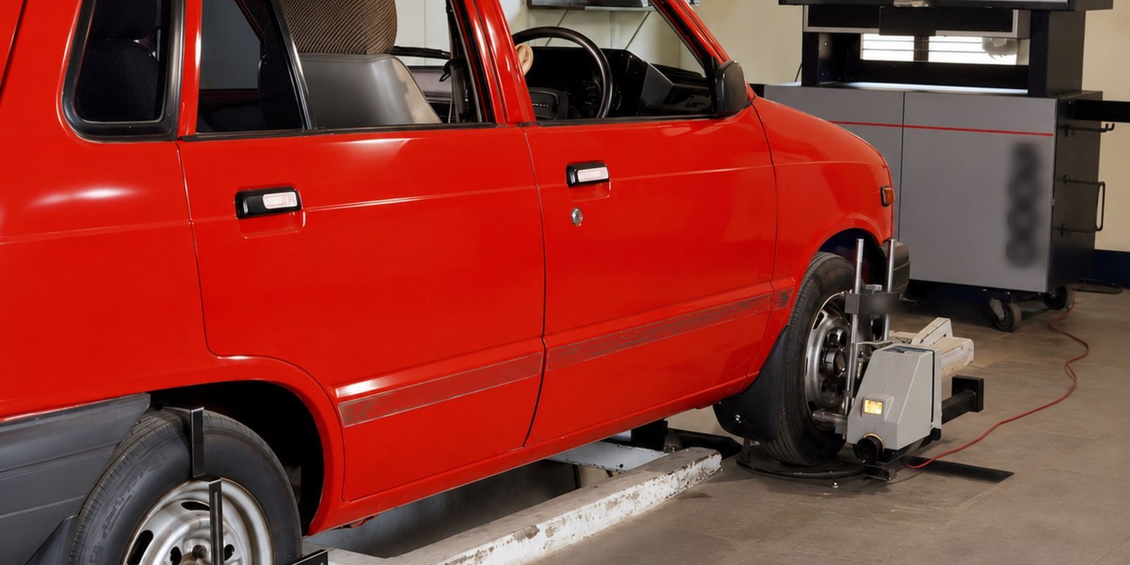 Carro vermelho sobre a plataforma de alinhamento de direção, com garras de medição presas à roda dianteira e o equipamento de leitura ao lado, numa oficina