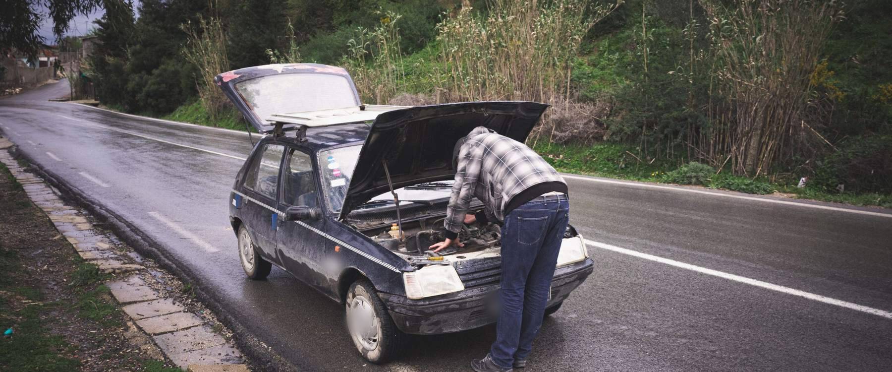 Homem de camisa xadrez inclinado sobre o motor de um carro pequeno e escuro, com capô e porta-malas abertos, parado no acostamento de uma estrada molhada junto a um barranco com vegetação
