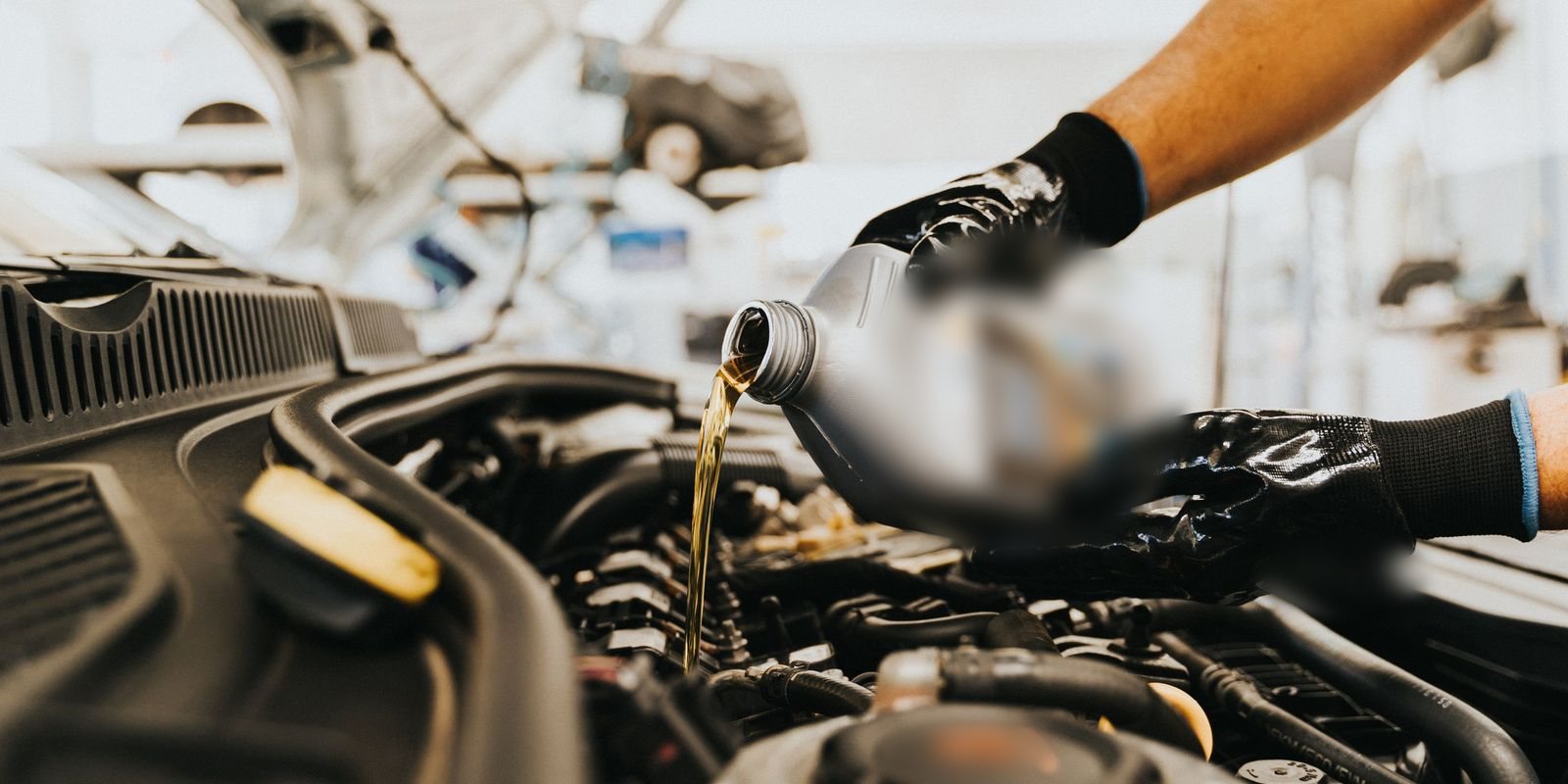 Mãos de luvas pretas despejam óleo dourado de um frasco no bocal do motor de um carro, numa oficina clara e desfocada ao fundo